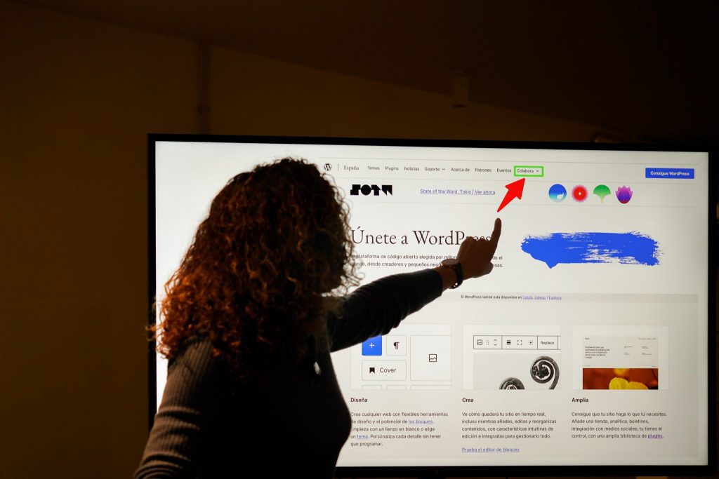 A person with curly hair is silhouetted against a large computer screen, pointing at a section of a webpage. The screen displays a website in Spanish, featuring the WordPress logo and navigation menu, with a red arrow pointing to the “Colabora” menu item.